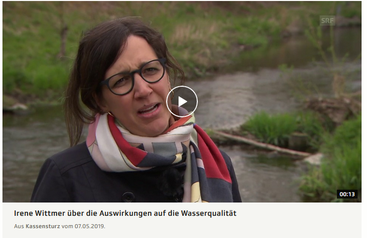 Eine Frau mit Schal steht neben einem Fluss und erzählt im Video über die Auswirkungen aus die Wasserqualität.