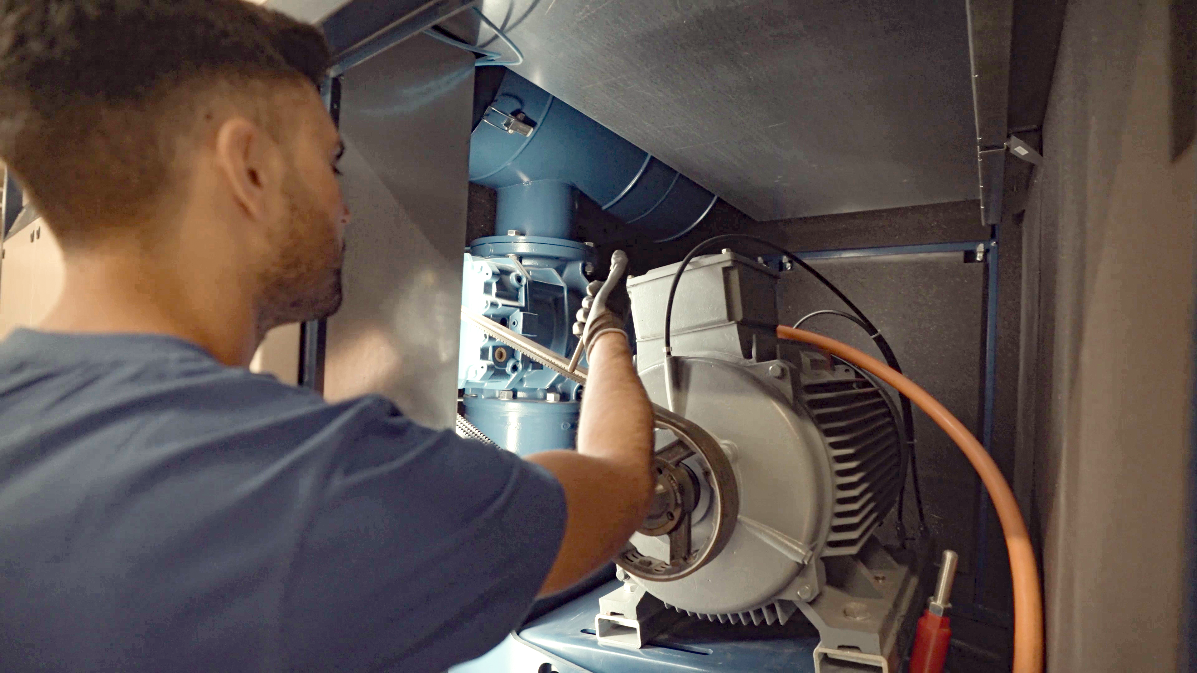 Eine Person im blauen Hemd inspiziert oder repariert Industriemaschinen innerhalb eines Metallgeh&auml;uses, wobei sie ein Werkzeug benutzt und Handschuhe tr&auml;gt.