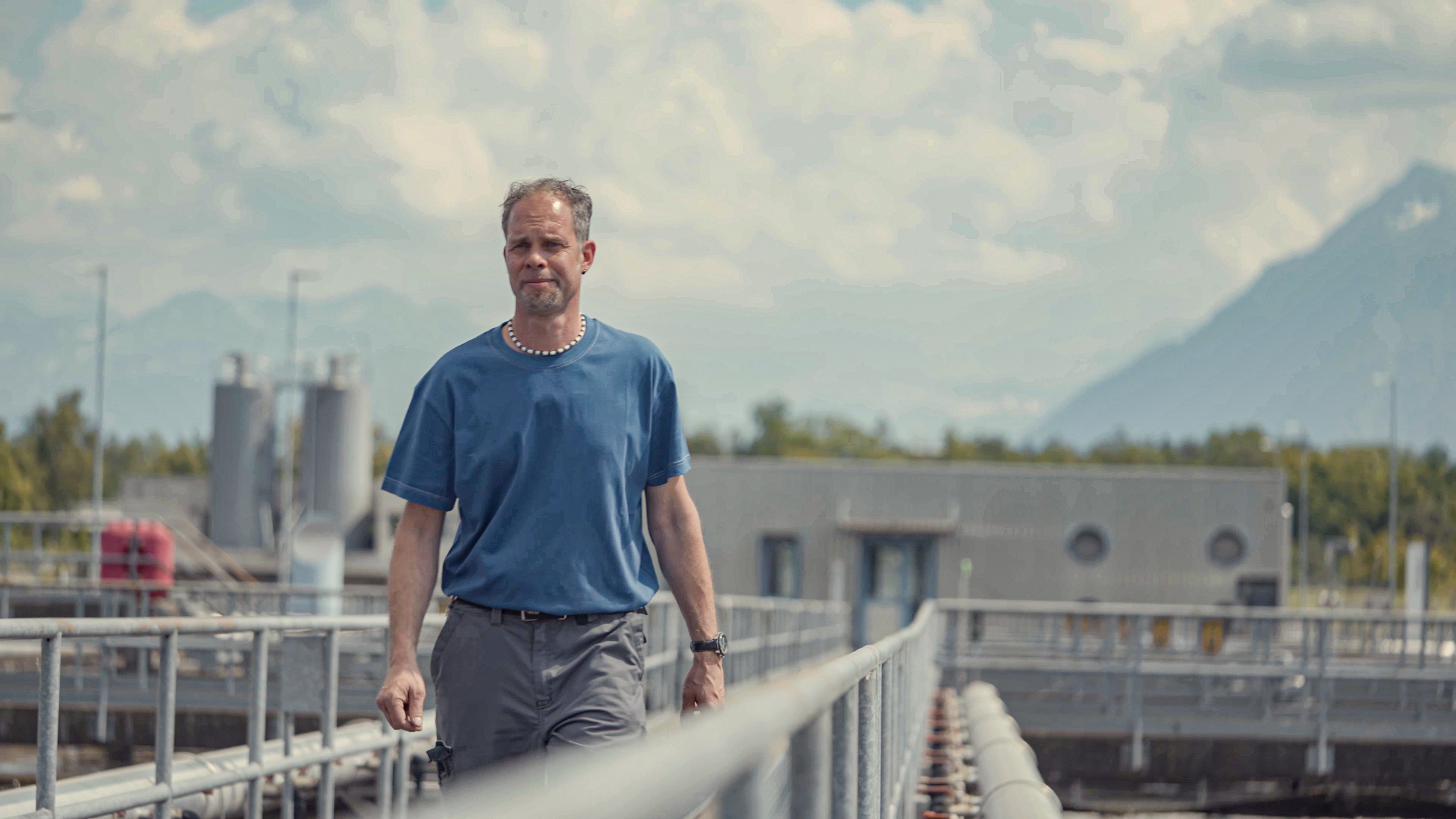 Ein Mann in einem blauen T-Shirt geht &uuml;ber einen Industrieweg mit Geb&auml;uden, Ger&auml;ten und Bergen im Hintergrund.
