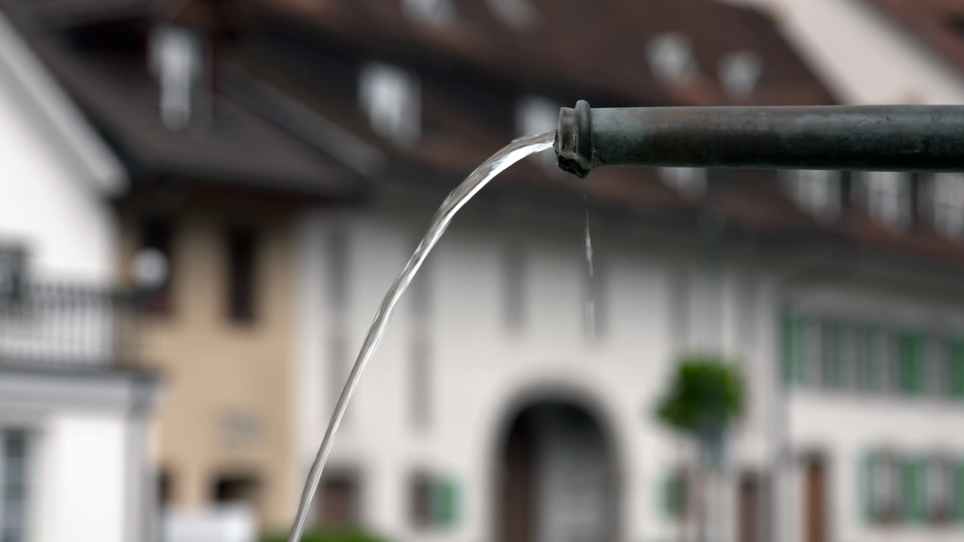 Aus einem Metallrohr str&ouml;mt ein gleichm&auml;&szlig;iger Wasserstrahl, im Hintergrund sind unscharfe Geb&auml;ude und Fenster zu sehen.
