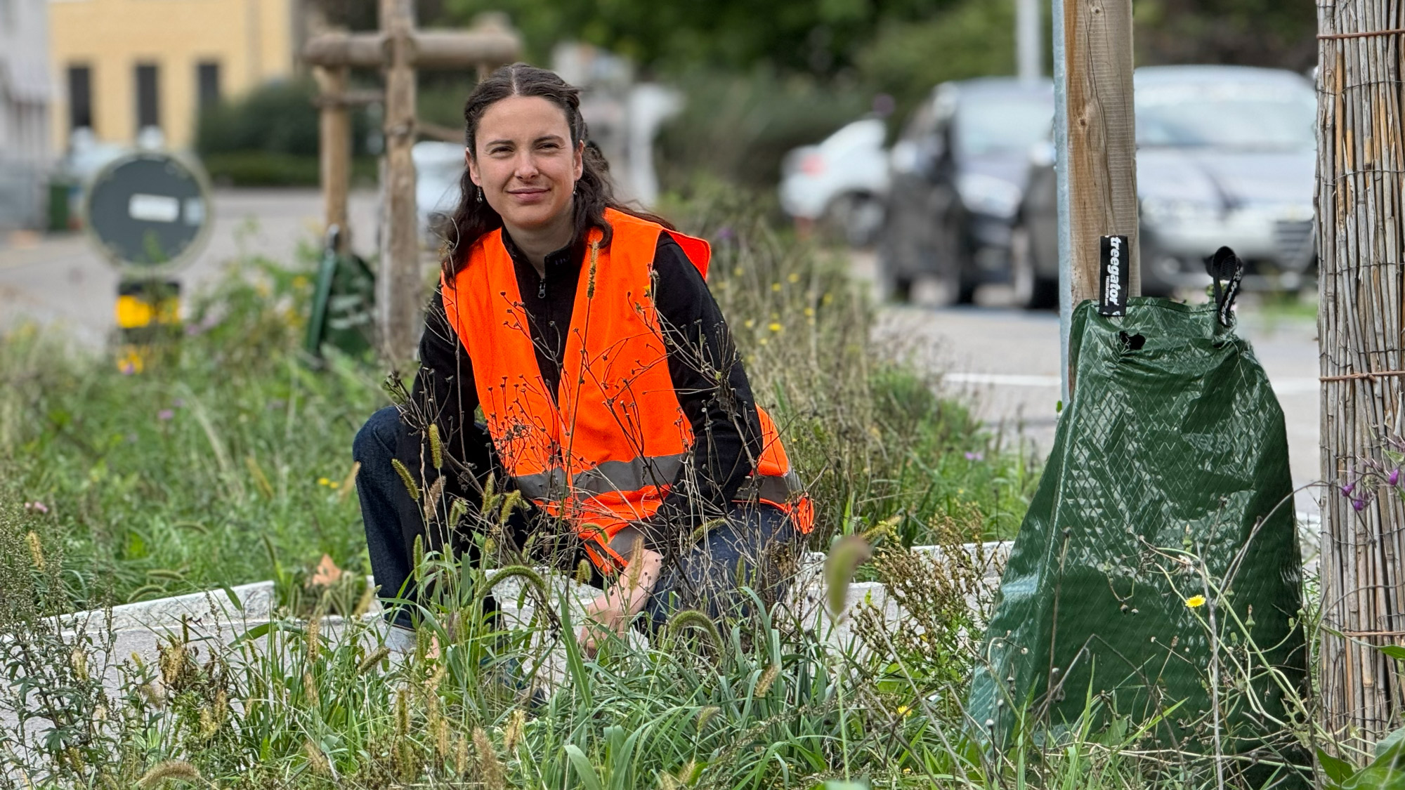 Eine Person, die eine orangefarbene Warnweste trägt, hockt in einem grasbewachsenen, städtischen Garten in der Nähe der Straße, mit geparkten Autos und Gebäuden im Hintergrund.
