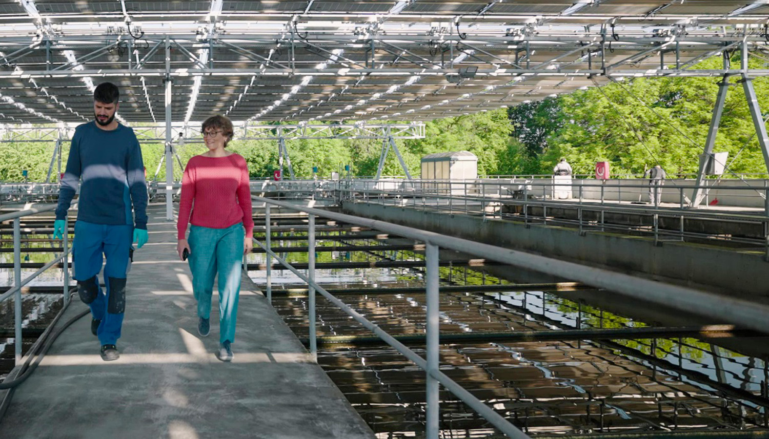 Zwei Personen gehen auf einem Betonsteg im Inneren einer großen Wasseraufbereitungsanlage mit Metallstrukturen und durch das Dach einfallendem Sonnenlicht.