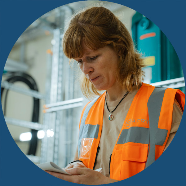 Eine Frau in einer orangefarbenen Sicherheitsweste schreibt in einer Industrieumgebung auf einem Notizblock, w&auml;hrend im Hintergrund Regale und Ger&auml;te zu sehen sind.