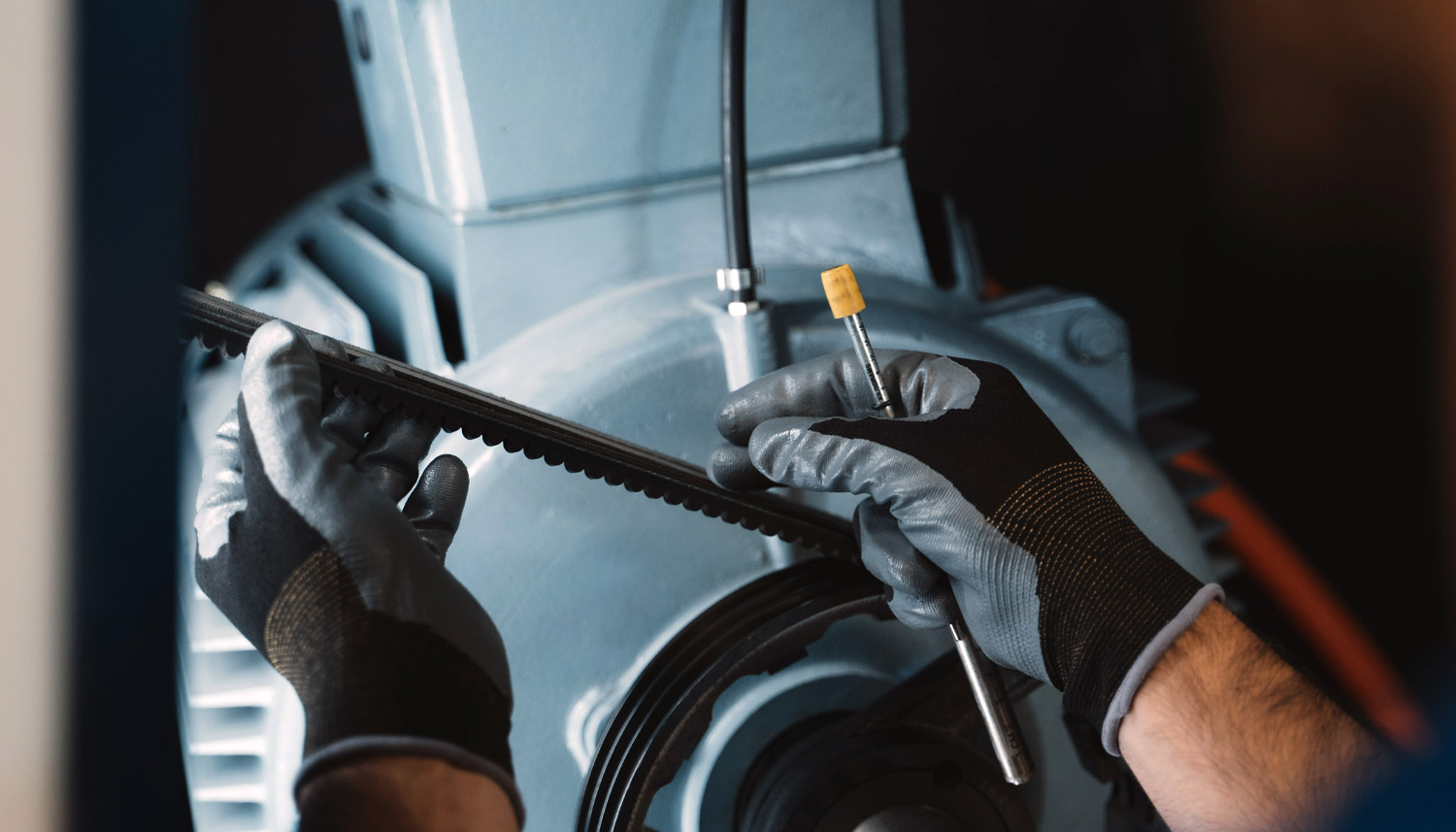 Eine Person mit Handschuhen f&uuml;hrt Wartungsarbeiten an Maschinen durch und h&auml;lt einen G&uuml;rtel und ein Werkzeug vor einen gro&szlig;en Industriemotor.