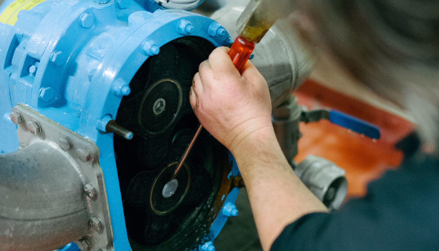Eine Person f&uuml;hrt mit einem Schraubenzieher Wartungsarbeiten an einer blauen Industriemaschine mit sichtbaren Zahnr&auml;dern und Metallrohren durch.