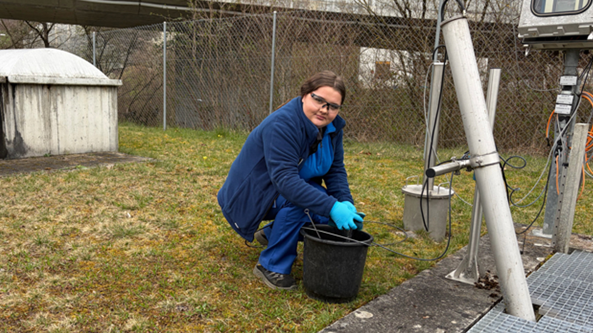 Eine Person in blauer Kleidung und mit Handschuhen entnimmt eine Probe mit einem schwarzen Eimer in der N&auml;he von wissenschaftlichen Ger&auml;ten auf einer Wiese.
