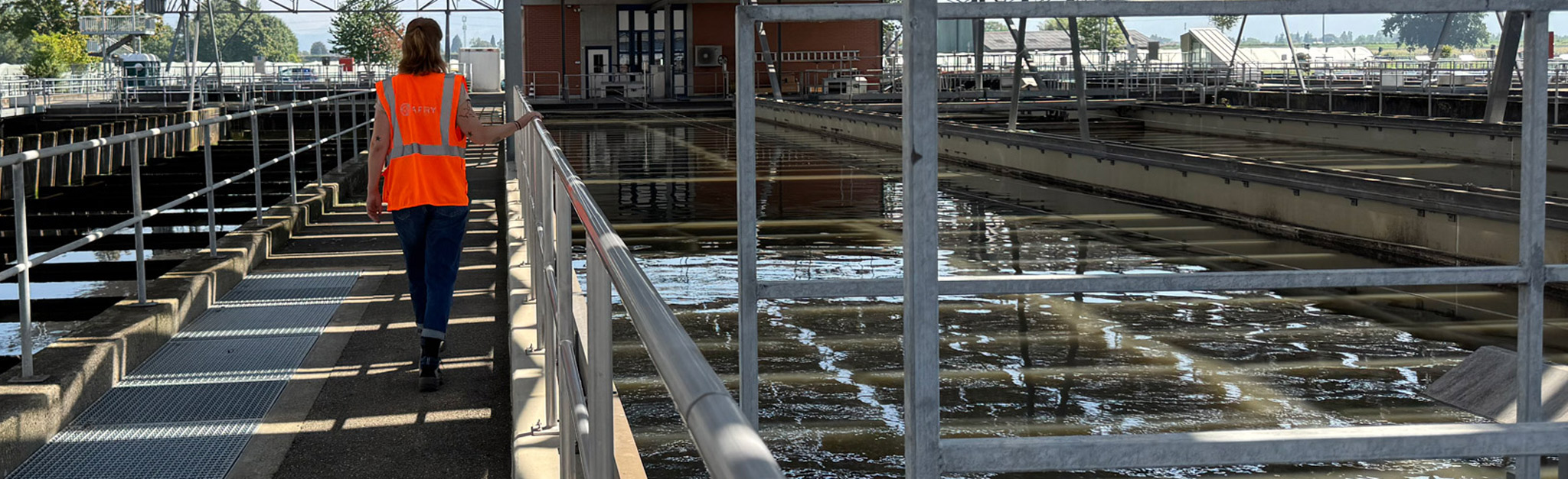 Eine Person in einer orangefarbenen Sicherheitsweste geht auf einem Gehweg neben großen rechteckigen Tanks in einer Wasseraufbereitungsanlage entlang.