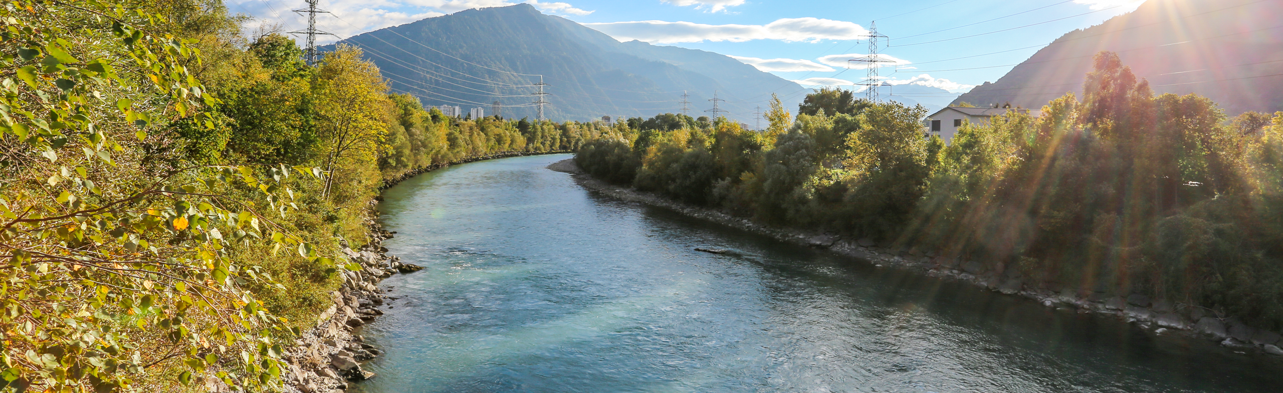 Ein Fluss schlängelt sich durch eine grüne Landschaft mit Bäumen auf beiden Seiten, Stromleitungen über dem Boden und Bergen im Hintergrund unter einem teilweise bewölkten Himmel.