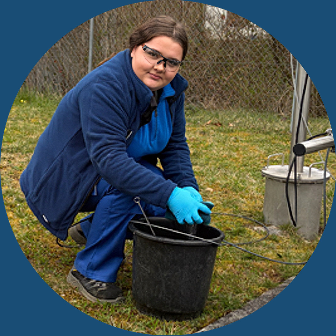 Eine Person mit blauer Kleidung und Handschuhen hockt im Gras und h&auml;lt einen schwarzen Eimer mit Ausr&uuml;stung in der N&auml;he eines Metallger&auml;ts im Freien, wahrscheinlich bei der Durchf&uuml;hrung eines Feldversuchs oder einer Wasserprobe.