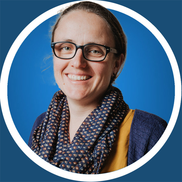 Eine Frau mit Brille, einem gemusterten Schal und einem gelben Oberteil l&auml;chelt vor einem blauen Hintergrund in die Kamera.