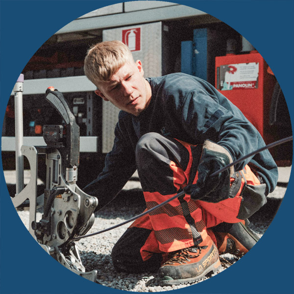 Ein Arbeiter in Schutzkleidung bedient im Freien Maschinen und hantiert mit Kabeln; im Hintergrund sind Arbeitsger&auml;te und ein Feuerl&ouml;scher zu sehen.