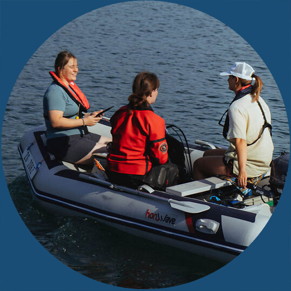 Drei Personen sitzen in einem Schlauchboot auf ruhigem Wasser, tragen Schwimmwesten und Freizeitkleidung und unterhalten sich.
